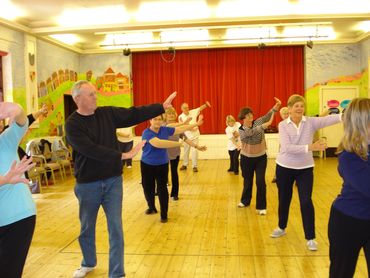 older white people are shown spaced out indoors following tai chi instructions from a tutor