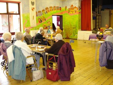 groups of older white people sit around small tables in groups listening to a bingo caller