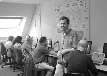 a black and white photo of a white man who is smiling at the camera while 4 people sit using compute
