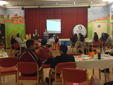 young people sit around tables facing a stage with a counselor giving a talk