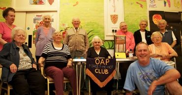 a group of older people sit and stand around a table which has a banner in the middle stating OAP Fo