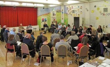 a group of people sit in a circle of chairs listening to a table of presenters