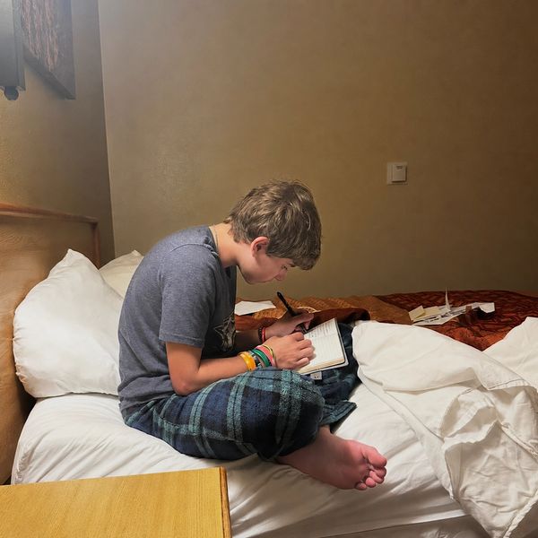 A boy in pajamas writing in a notebook while sitting on a bed.