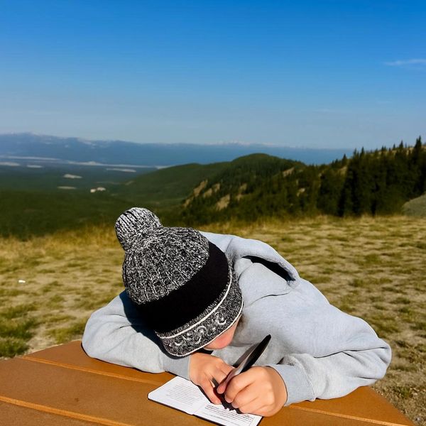 Person writing in a notebook outdoors with mountains in the background.