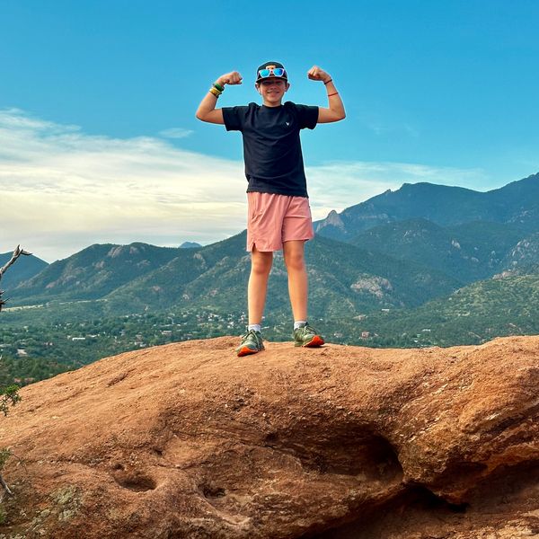 Boy flexing muscles on a rocky hill with mountains in the background.