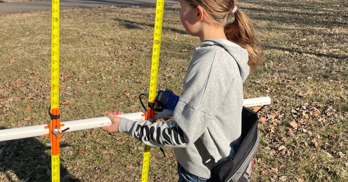A girl measures a white pipe between two yellow measuring sticks outside.