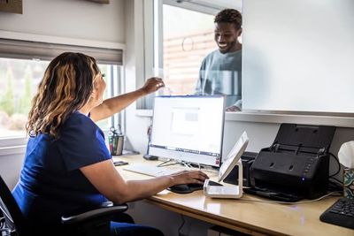 Receptionist assisting a smiling visitor through a glass window.