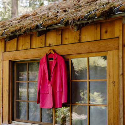 A hot pink blazer hanging outside a wood cabin with a mossy roof. The photograph was taken by Krysta