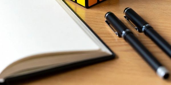 A solved Rubik's Cube on a wooden desk with pens and a notebook.