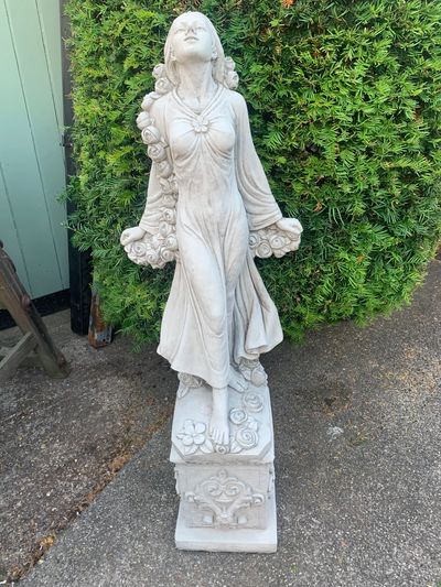White stone statue of a woman holding flowers, standing on a decorative pedestal.