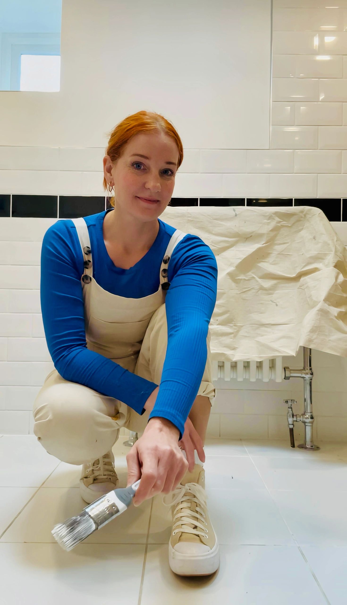 Woman in blue shirt painting white tiles on floor with a brush.