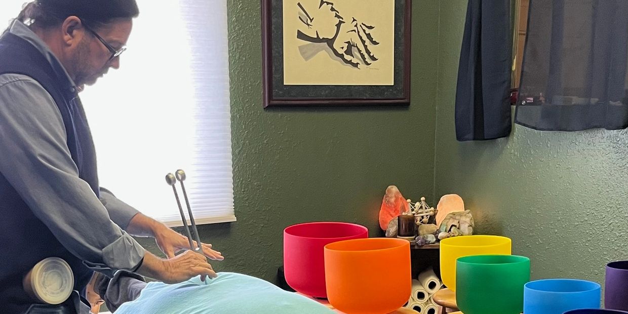 A man uses tuning forks near a person lying down with colorful bowls nearby.