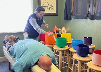 A man plays colorful singing bowls near a person lying on a massage table.