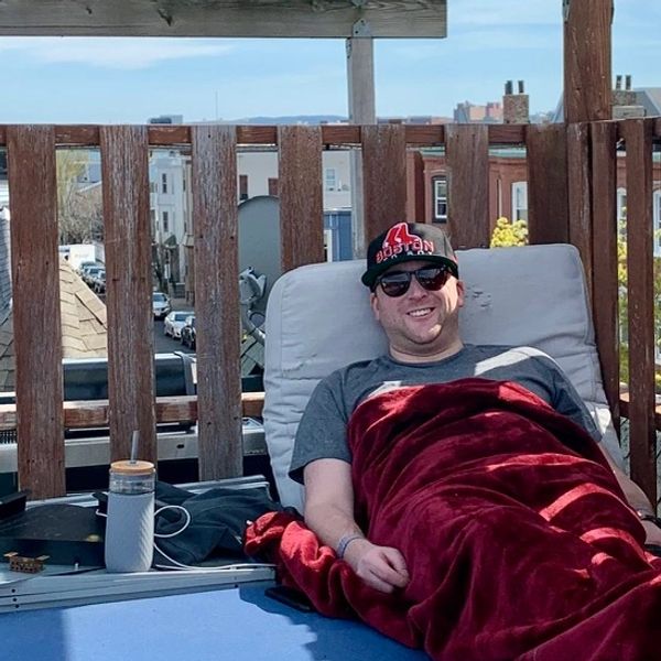 Man relaxing outdoors on a chair wrapped in a red blanket, wearing sunglasses and a Boston cap.