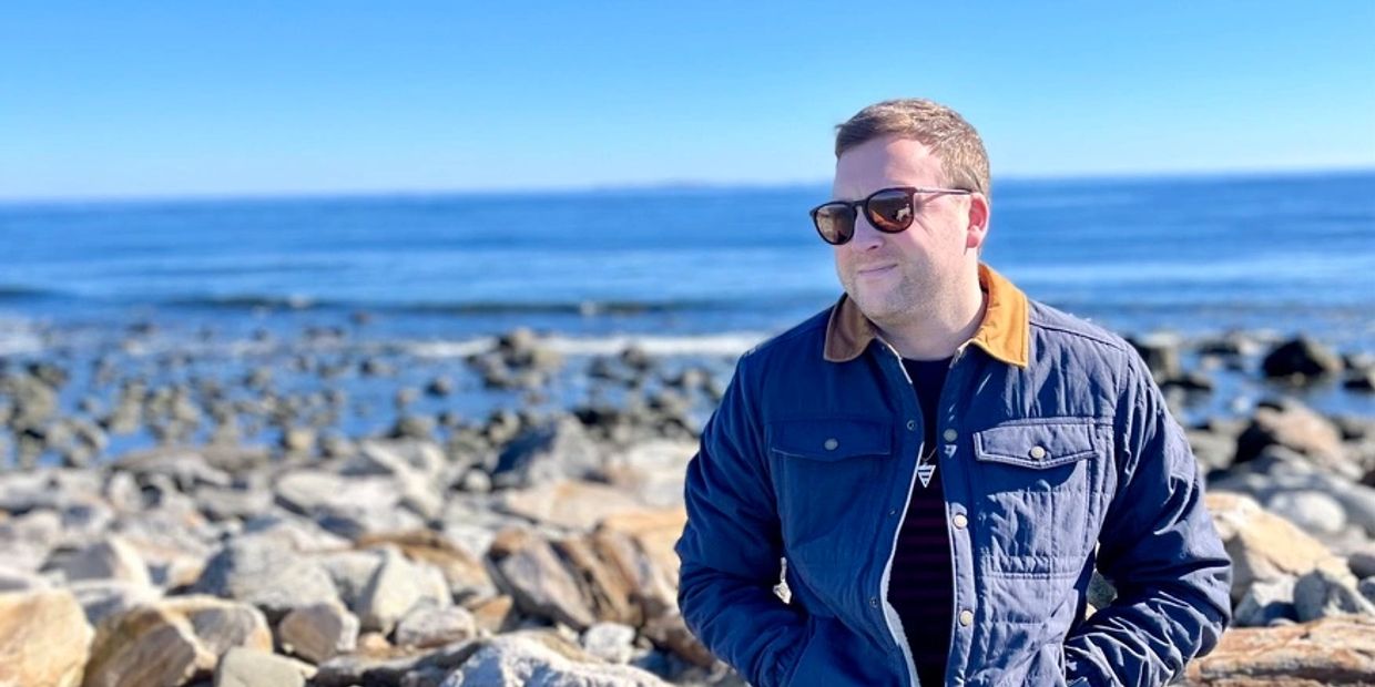 Man wearing sunglasses and a blue jacket stands by rocky seaside under clear blue sky.