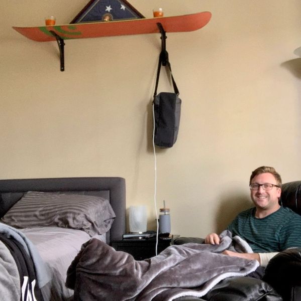 Man relaxing on a couch with a gray blanket, beside a bed and nightstand.