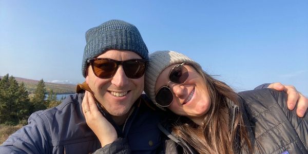 A smiling couple wearing sunglasses and winter hats outdoors on a sunny day.