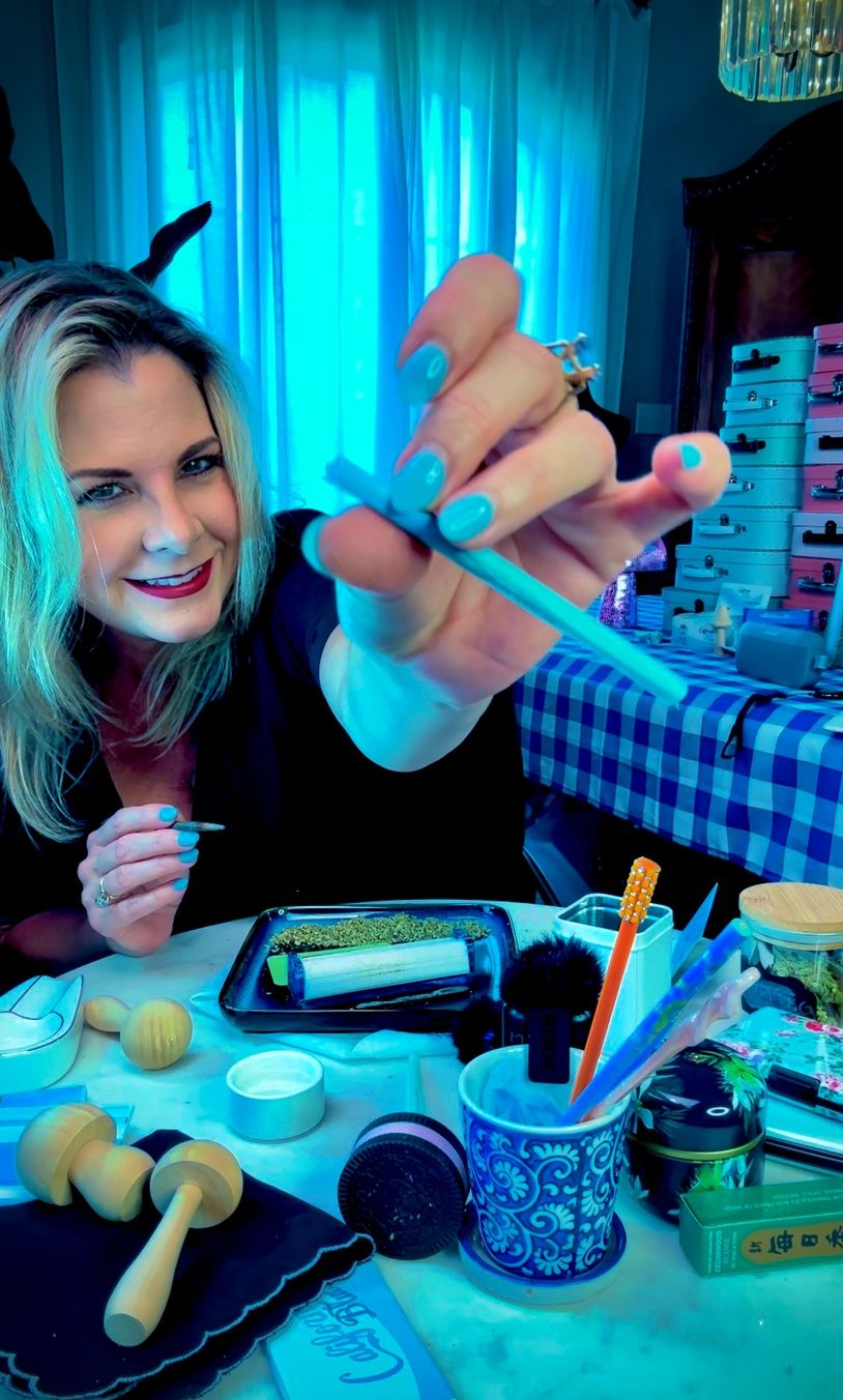 Woman smiling and holding a rolled joint at a cluttered table with smoking accessories.