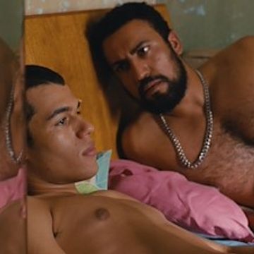 Two shirtless men lying close together, one looking intently at the other.