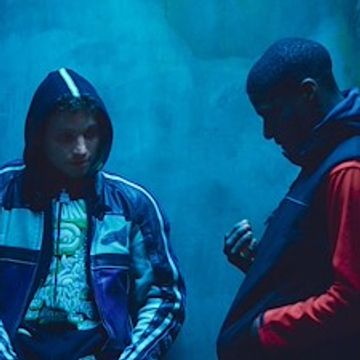 Two young men in hoodies stand in a dimly lit blue room, looking contemplative.