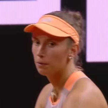 Female tennis player in an orange visor and outfit focused during a match.