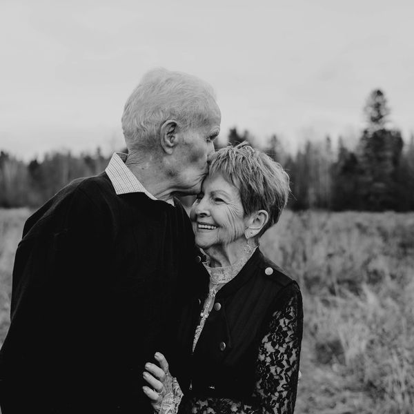 Elderly man tenderly kisses woman on forehead in a serene outdoor setting.