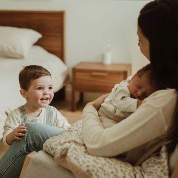 A joyful toddler looks at a woman holding a newborn baby in a cozy bedroom.