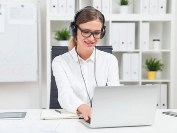 Smiling woman wearing headphones working on a laptop in a bright office.