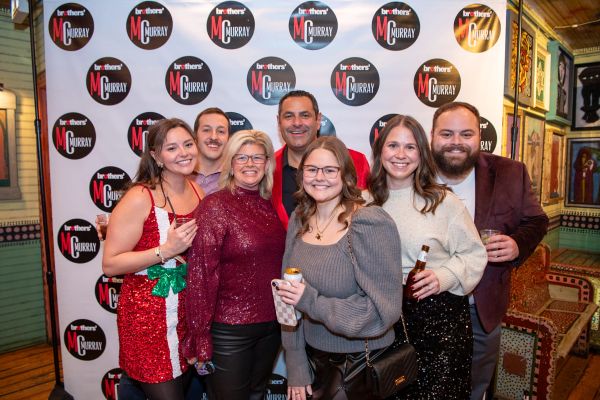 Group of seven smiling friends at a festive event with Brothers MC Murray backdrop.