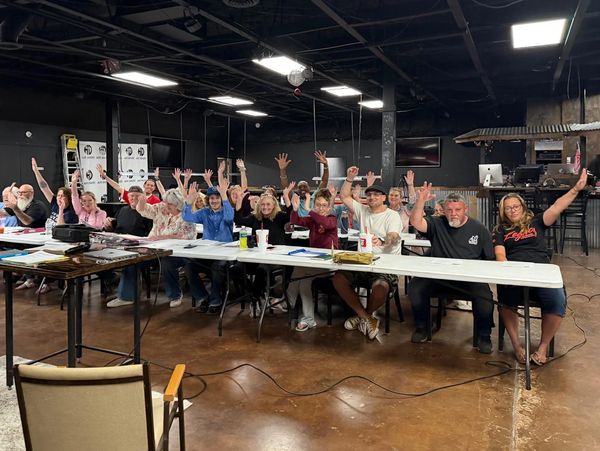 A group of people sitting at tables raising their hands in a meeting.