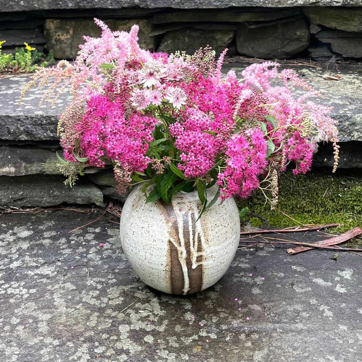 A rustic vase filled with vibrant pink and purple flowers against a stone backdrop.