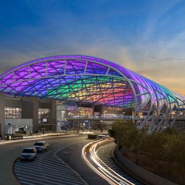 A colorful illuminated canopy covers a modern airport terminal entrance at dusk.