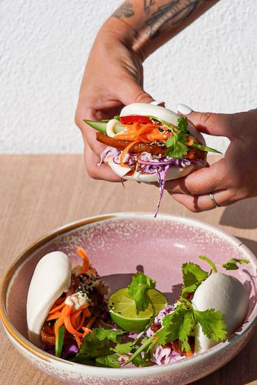 Hands holding a veggie bao bun over a bowl with more buns and lime.