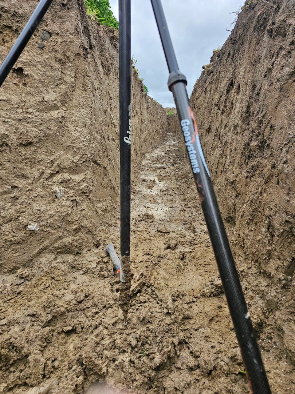 Surveying poles leaning against the soil wall of an excavated trench.
