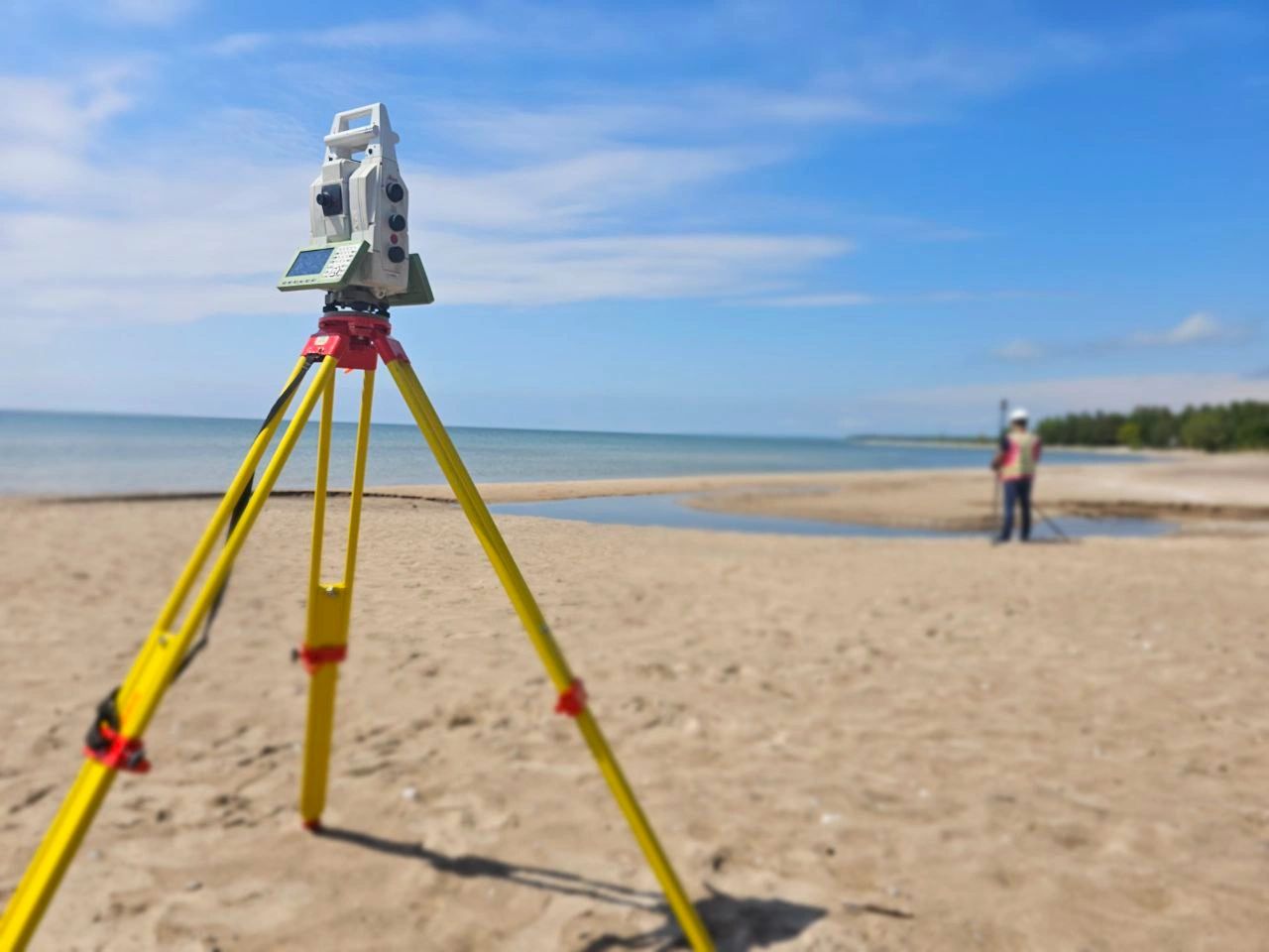 Total station on a yellow tripod at a sandy shoreline, with a surveyor working near the water.