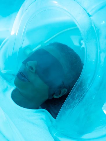 Person with eyes closed inside a transparent chamber emitting blue light.