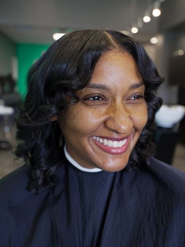 A woman with styled wavy hair smiles brightly in a salon.