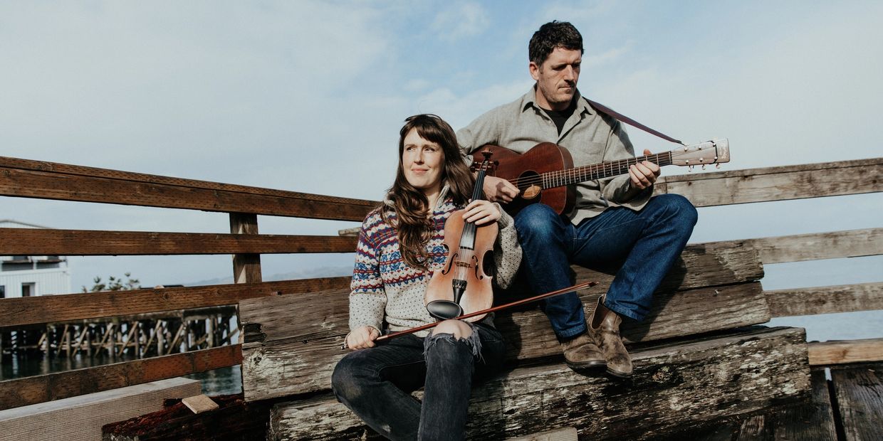 Two musicians playing violin and guitar outdoors on wooden benches.