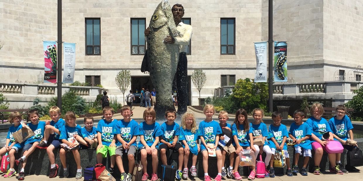 Group of children in blue t-shirts posing in front of a large fish statue.