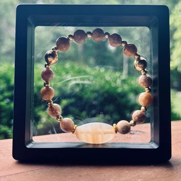 Wooden beaded bracelet displayed in a black square frame with a blurred green background.