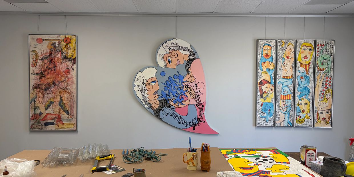Colorful abstract paintings displayed on a white wall above a cluttered work table.