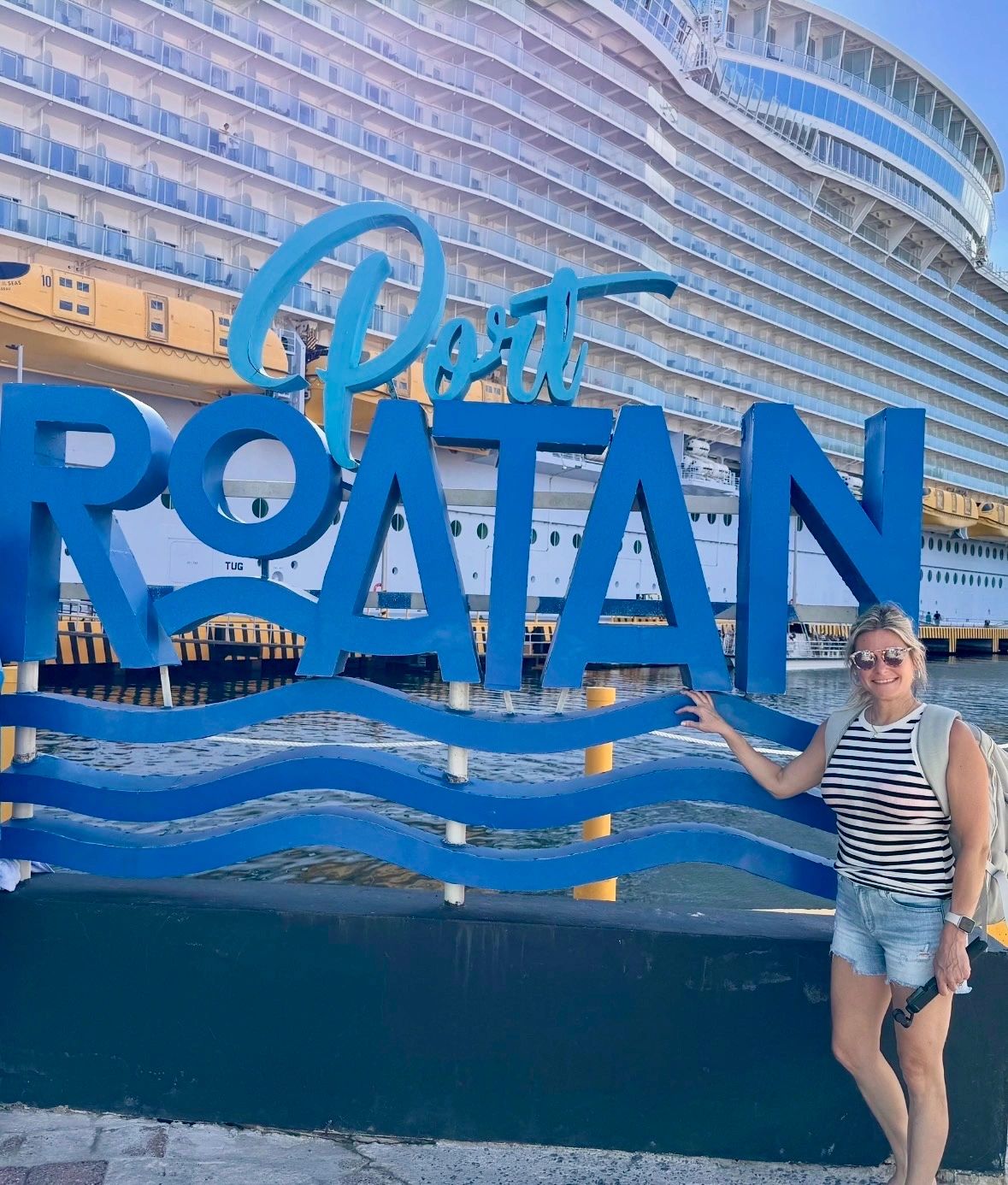 Woman posing by the 'Port Roatan' sign with a cruise ship in the background.