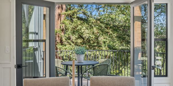 Balcony with a table and chairs overlooking lush green trees through open glass doors.