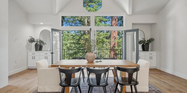 Bright dining room with wooden table, mixed chairs, and large open windows showing green trees.