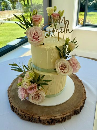 Two-tiered elegant wedding cake adorned with pink roses and greenery.