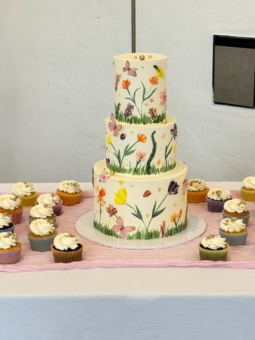 Three-tiered floral cake surrounded by cupcakes on a table.