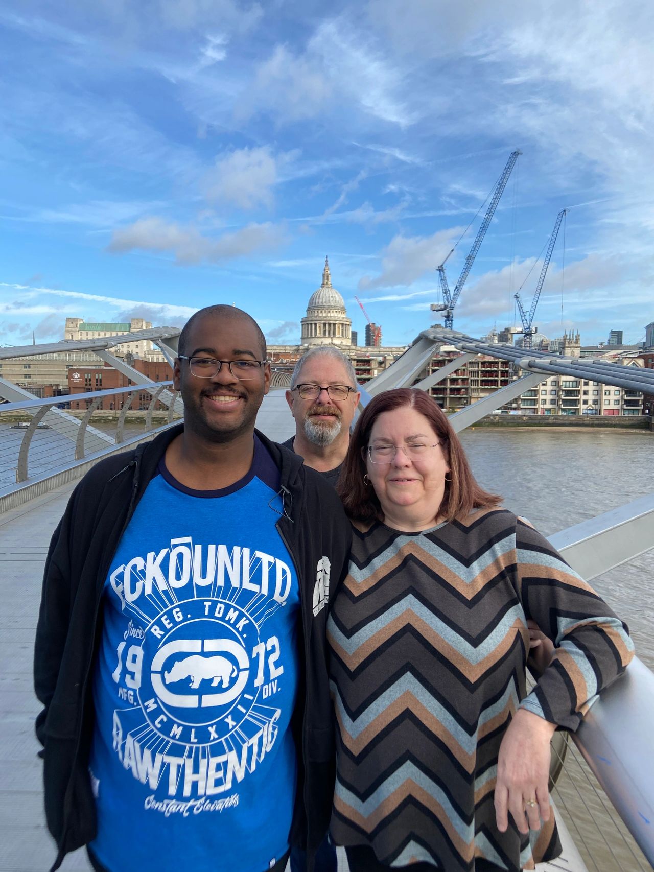 A group photo of three people standing on the Millennium Bridge in London with the iconic dome of St. Paul's Cathedral and the Thames riverfront in the background, a key filming location for the Death Eater attack in Harry Potter.