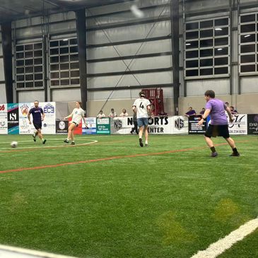 Adults playing indoor soccer on artificial turf in a sports center.