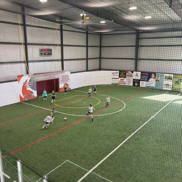 Indoor soccer game in progress on artificial turf with players and a goalkeeper.