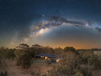 Milky Way arching over a cozy house in a forest clearing at night.